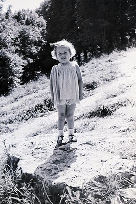 Young girl in a dress and Mary Jane shoes stands on a rocky, grassy slope. Appears to be mid-1940s, based on clothing style a...