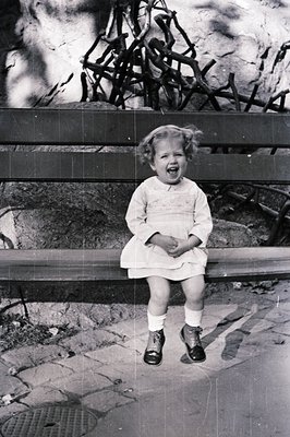 A young girl in a collared dress and Mary Jane shoes sits on a park bench, laughing broadly. Background shows foliage & a scu...