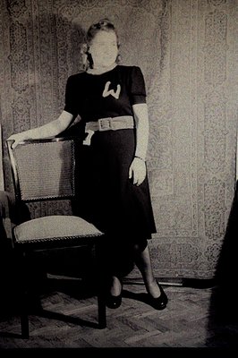 A woman in a dark dress with a wide belt poses leaning on a wicker chair. The dress features a large, stylized "W" emblem on ...