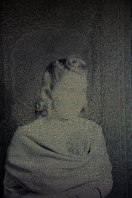 A heavily pixelated, monochrome portrait of a person seated and appearing draped in fabric, possibly a shawl or sweater. The ...