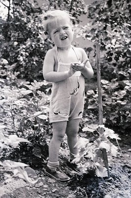 A toddler wearing denim overalls and dark shoes stands outdoors amidst lush foliage, appearing excited with open palms. The p...