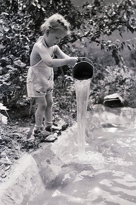 Young girl in a short romper plays at a natural spring or stream. She carefully pours water from a metal pail. The scene capt...