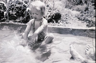 A young child, in a patterned swimsuit, sits in shallow water, playfully splashing. Visible are the adult's feet beside the c...