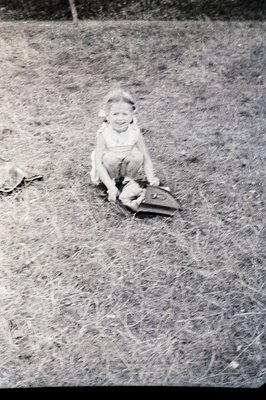 A young child, appearing to be a boy, sits on a frisbee in a grassy field. Visible through the foliage behind is a suggestion...