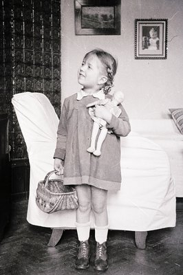 A young girl in a collared dress, knee-high socks, and boots stands holding a doll and a woven basket. Set against a decorati...