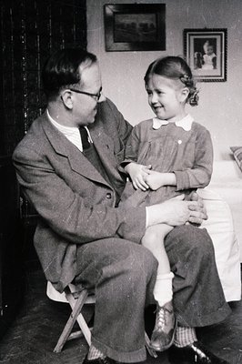 Man in a suit and glasses sits on a folding chair, gently holding a young girl in a dress. Interior scene with framed artwork...