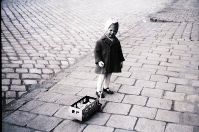Young child, dressed in a wool coat and tights, stands with a small wagon on a cobblestone street. Appears to be outdoors, li...