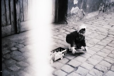 A young child, wearing a cap, playfully pulls a wooden toy cart across a stone-paved surface. The scene is likely outdoors, w...