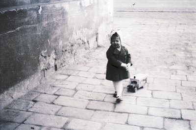 A young girl in a dark coat and cap walks along a stone-paved area next to a textured wall. She carries a small, square box. ...
