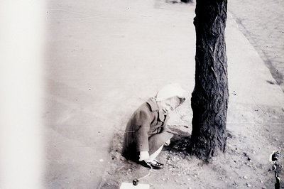 A young child in a distinctive cap and coat crouches near a tree trunk, intently focused on something in their lap—perhaps dr...