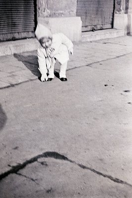 A young child in a formal white suit and hat kneels on a concrete surface, appearing to reach for something. A closed shutter...