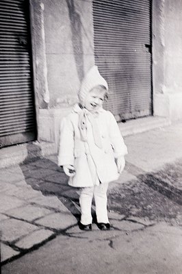 A young child, bundled in a white coat and hat, stands outdoors near a closed storefront. The child’s expression is joyful, w...