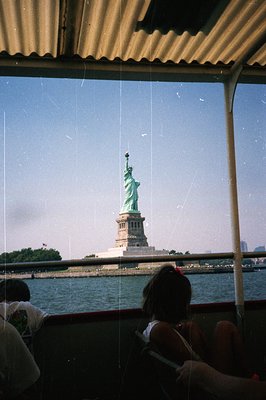Framed by a boat's canopy, the Statue of Liberty rises from the harbor, seen over water. A hint of Manhattan’s skyline is vis...