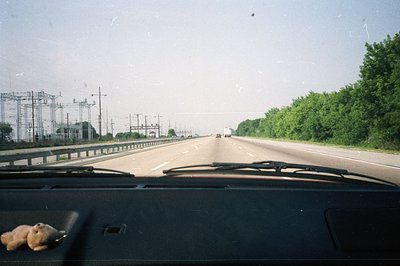 A point-of-view shot from inside a vehicle on a multilane highway. Power lines and a substation dominate the left background,...