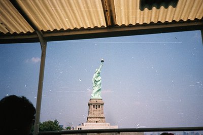 View of the Statue of Liberty framed by a corrugated roof, likely taken from a ferry or observation point. The statue’s green...