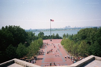 High-angle view of a crowd gathered around a large American flag at Liberty State Park, NJ. A sprawling plaza of brick paving...