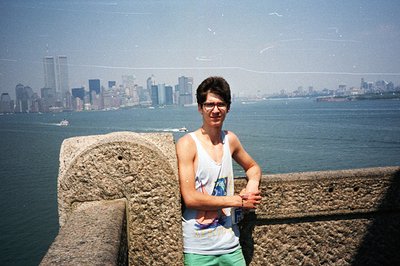 A young man in a tank top and shorts poses near a stone parapet overlooking the New York City skyline, including the World Tr...