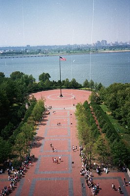 Aerial view of a memorial plaza with a large American flag, overlooking a harbor & distant city skyline. Brick paving, manicu...