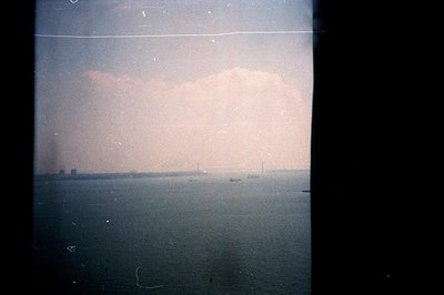 A hazy, coastal cityscape viewed across a body of water. Distant industrial buildings and a few ships are visible through den...