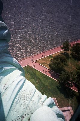 Close-up perspective of the Statue of Liberty’s robes overlooking a grassy area and waterfront walkway. Tourists are visible ...