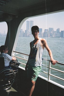 A young man in a tank top and shorts stands on a ferry, the New York City skyline visible in the background. A seated individ...