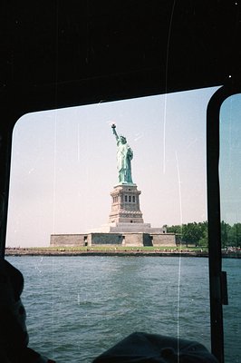 A view of the Statue of Liberty, seen through a window frame. The statue stands on a pedestal against a clear blue sky and wa...
