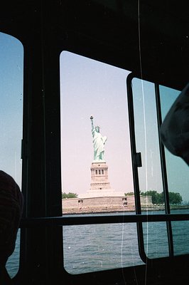 View of the Statue of Liberty framed within a ship's porthole window. The monument stands on a pedestal, overlooking water wi...