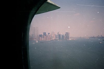 View from an airplane window overlooking New York City skyline across a body of water. Visible are the World Trade Center tow...