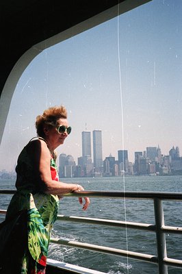A woman in a tropical print dress stands on a boat deck, gazing towards a New York City skyline featuring the iconic Twin Tow...