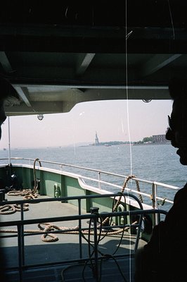 A ferry deck scene: a silhouetted figure gazes towards the distant Statue of Liberty framed by the ship’s railing and overhea...