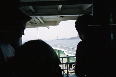 Two figures silhouetted against a view of the Statue of Liberty across the water. The photograph appears to be taken from ins...