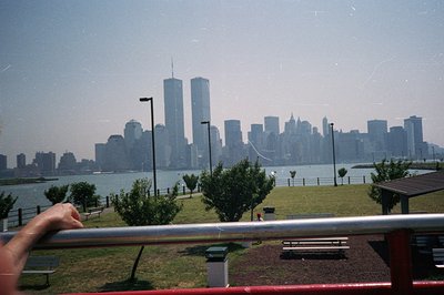 Panoramic view of the Manhattan skyline, dominated by the World Trade Center towers, across a body of water. Green lawn, tree...