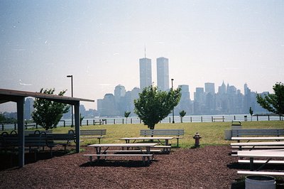 A picnic area faces a distant view of the Manhattan skyline, dominated by the iconic World Trade Center towers. The park feat...