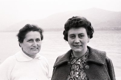 Two women, likely mother & daughter, pose outdoors with a coastal landscape behind them. Both wear vintage hairstyles and clo...