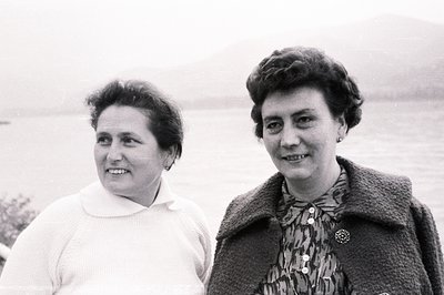 Two women with short, styled hair smile warmly, posed outdoors with a coastal view. The woman on the left wears a white sweat...
