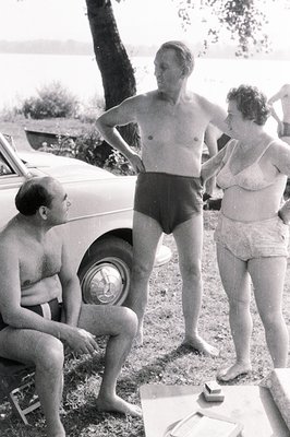 Black and white photo captures a lakeside scene in the 1960s. Two adults, a man seated & another standing, are dressed in vin...
