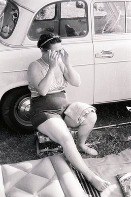 A woman in a vintage swimsuit and pearl necklace sits perched on a car’s tire, shielding her eyes. She appears to be outdoors...