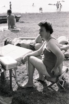 A woman sits on a folding chair in a grassy area near a beach, observing sunbathers. A picnic table with a checkered cloth si...
