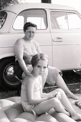 A young girl and woman pose outside a vintage car, likely a 1950s model. The girl wears a one-piece bathing suit, while the w...