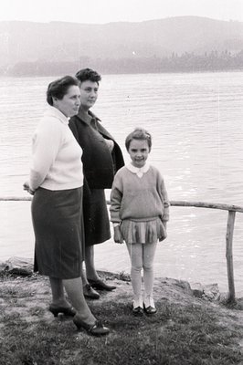 A young girl stands between two women near a body of water, possibly a lake. The women are formally dressed; the pregnant wom...