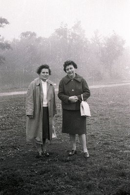 Two women in mid-length skirts and coats stand outdoors, appearing relaxed and engaged in conversation. Likely a casual portr...