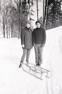 Two young men stand on a snow-covered slope, one wearing a dark coat and the other a dark turtleneck sweater. A vintage woode...