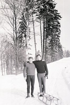 Two men stand proudly on snow, beside a wooden toboggan. Both wear dark sweaters and trousers, typical winter attire of the m...