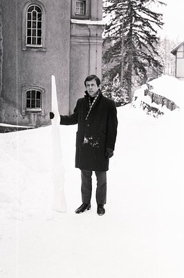 Man in a dark overcoat and turtleneck stands beside a snowdrift, holding a long, thin white object. Architectural backdrop su...