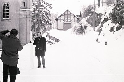 Black and white image shows a photographer capturing a man standing in a snow-covered courtyard. Traditional timber-framed bu...