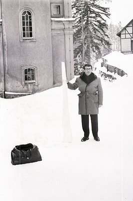 Man in a belted coat and glasses stands amidst deep snow, holding a thin, rectangular object aloft. A sturdy travel bag sits ...