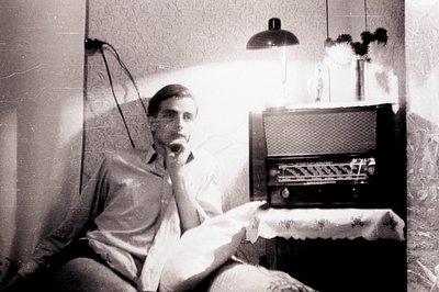 Young man seated, hand to chin, looking towards a vintage radio on a draped table. Stark lighting highlights textured walls &...
