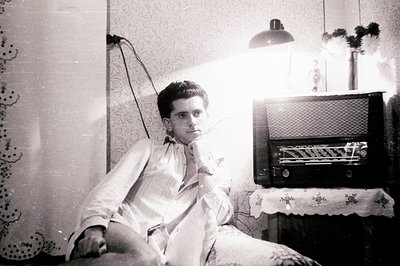 Young man in a relaxed pose, seated on a cushioned chair, framed by vintage wallpaper and a large, ornate radio. Likely 1950s...