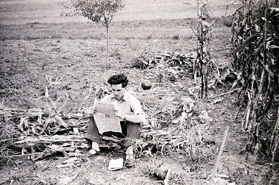 A man sits amidst tall corn stalks and debris, reading a newspaper. Appears to be outdoors, likely rural setting. The photogr...