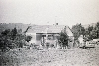 Classic rural scene: a modest, gable-roofed house with a prominent chimney & small windows, set within a fenced yard. A cross...